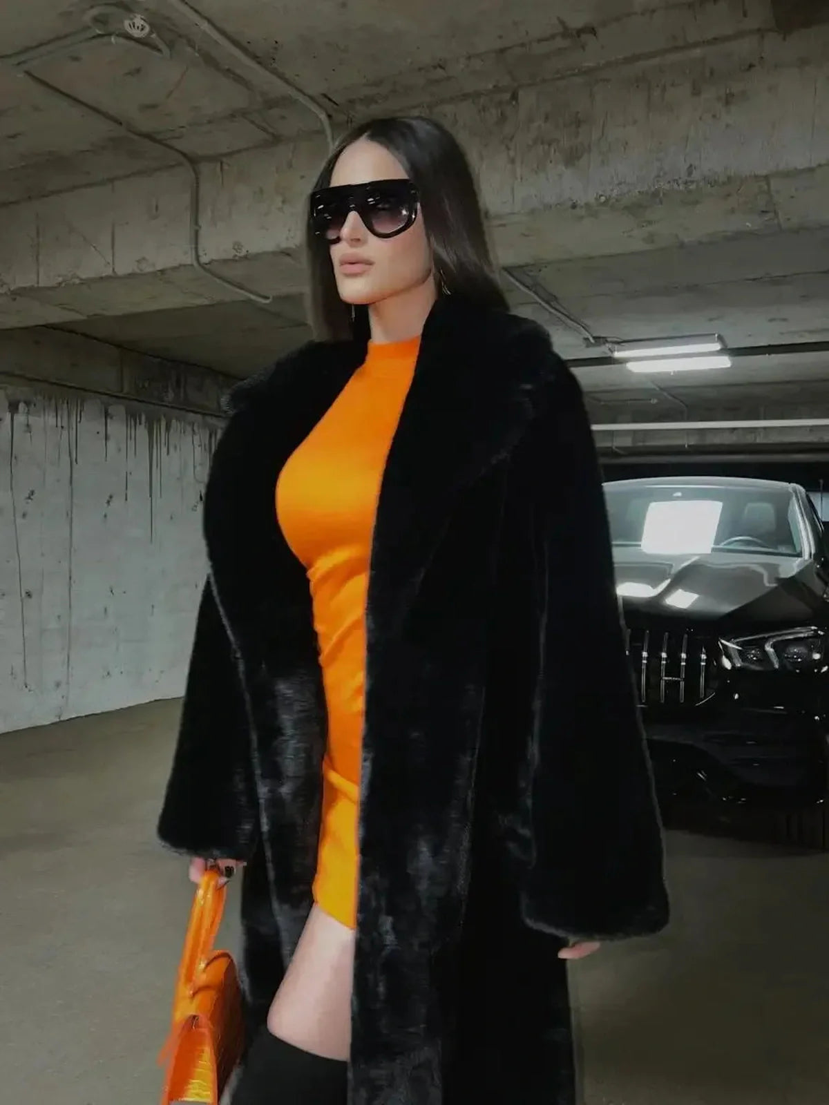 Woman in a stylish orange mini dress and black fur coat, accessorized with sunglasses and orange handbag.