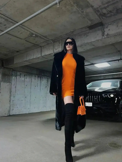 Stylish woman in an orange mini dress and black coat, showcasing a chic urban look in a parking garage.
