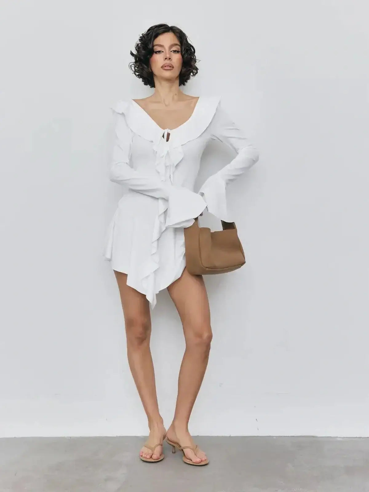 Model wearing a white mini dress with ruffles and a V-neckline, paired with a brown bag and sandals.