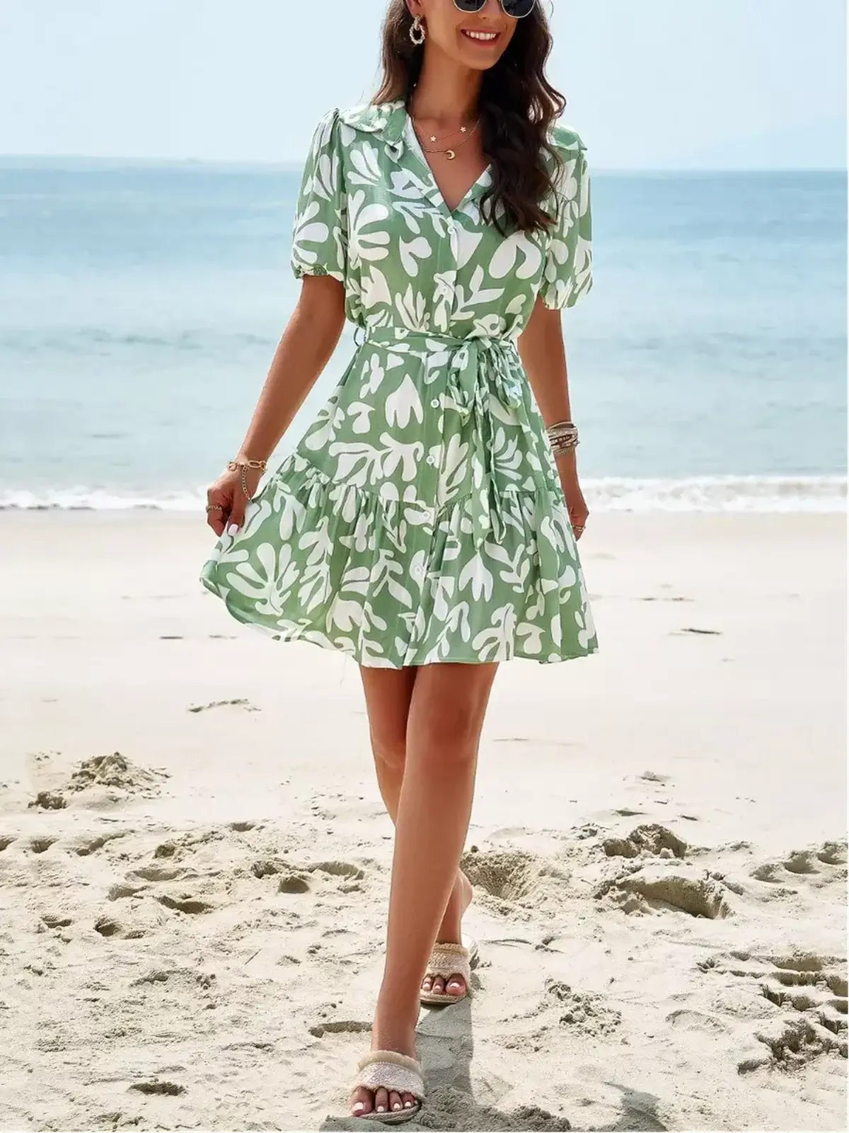 Model wearing Tropica Breeze Mini Dress, a short sleeve green floral dress, on the beach, ideal for summer parties.